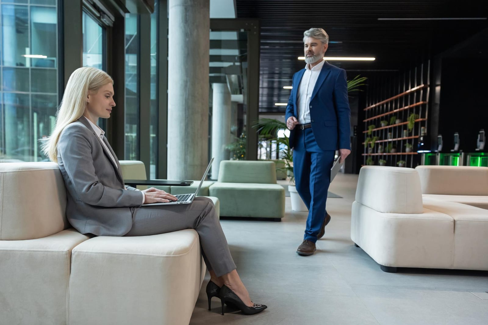 Eine Frau arbeitet auf einem Sofa in einer Lobby am Laptop, während ein Mann im blauen Anzug vorbeigeht.